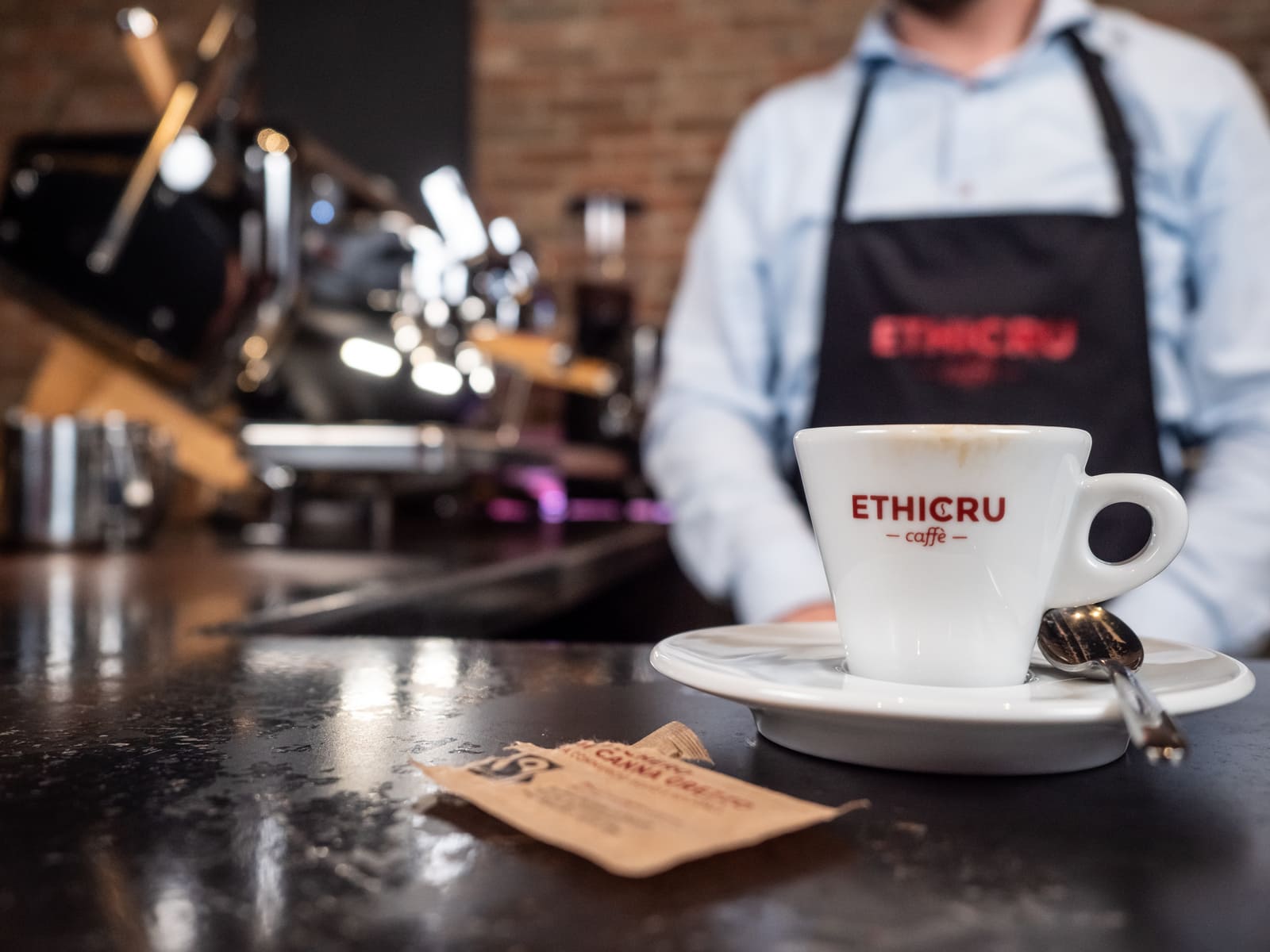 Barista con grembiule Ethicru serve un caffè in tazzina con logo Ethicru su bancone, con una bustina di zucchero biologico visibile.