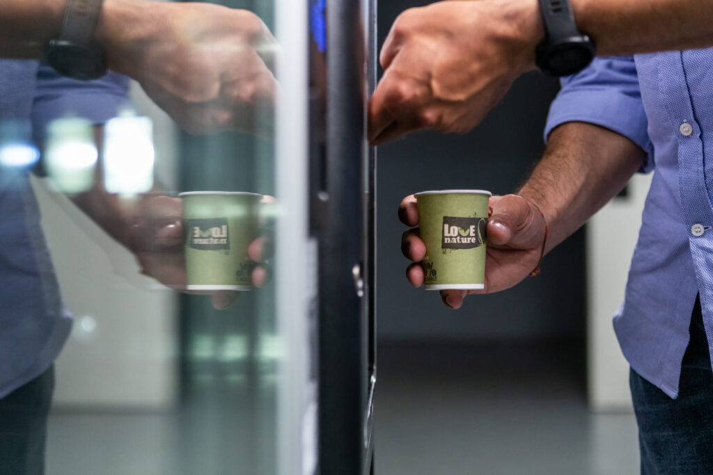 Main d'une personne prenant un verre de café dans un distributeur automatique, avec un gobelet compostable 100% biodégradable « Love Nature » reflété sur le verre.