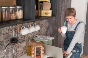 Donna con salopette in cucina con tazza di caffè in mano mentre utilizza un computer portatile, circondata da barattoli di cibo e utensili da cucina