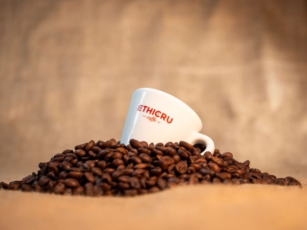 Tasse à café de la marque Ethicru posée sur un tas de grains de café torréfiés, sur un fond beige naturel, évoquant l'authenticité du café issu du commerce équitable.