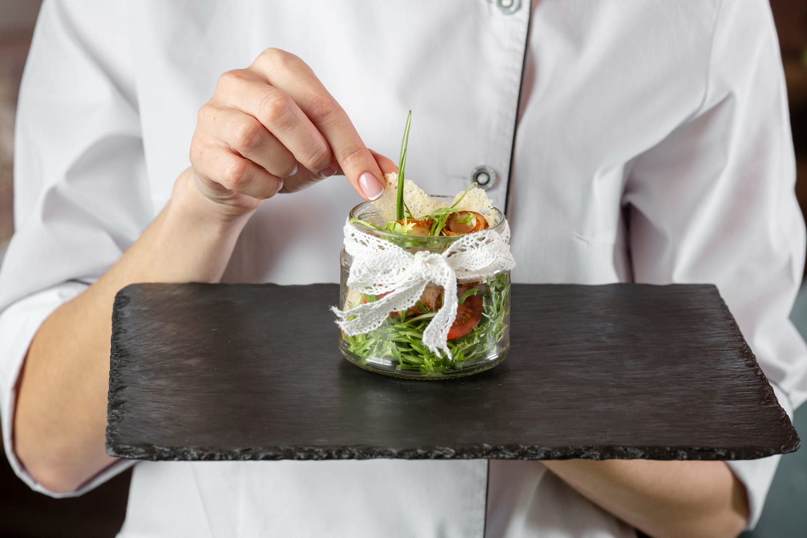 Insalata gourmet servita in un vasetto di vetro decorato con fiocco in pizzo, con verdure fresche e crostini, presentata su un vassoio di ardesia per un tocco elegante.
